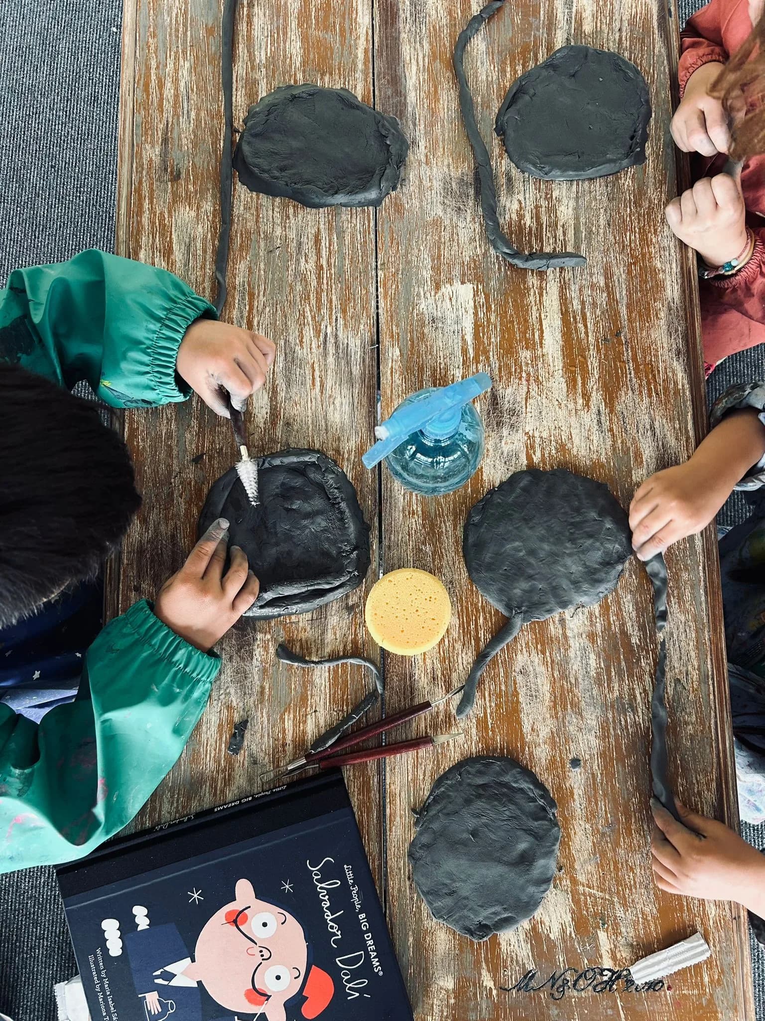 Children sculpting black clay slabs at a wooden table alongside a Salvador Dalí book