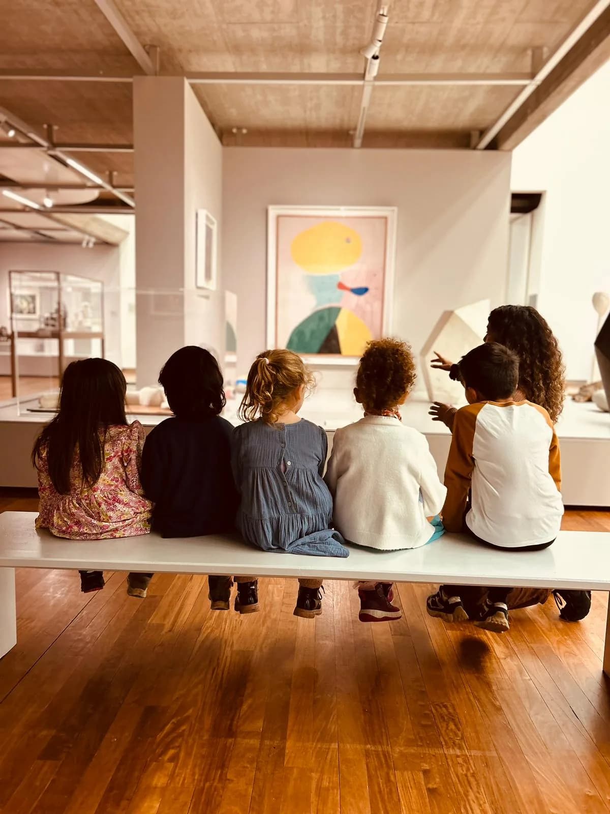 Six children sitting on a bench in a museum, looking at a large Miró-style painting