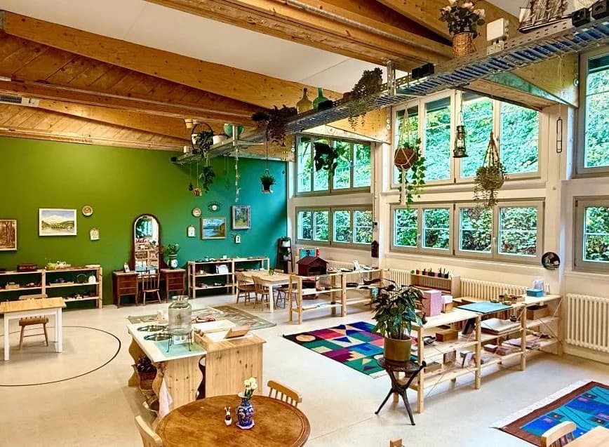Trinity Montessori — der helle grüne Primarklassenraum mit hohen Holzbalken, Pflanzen und vorbereiteten Montessori-Materialien